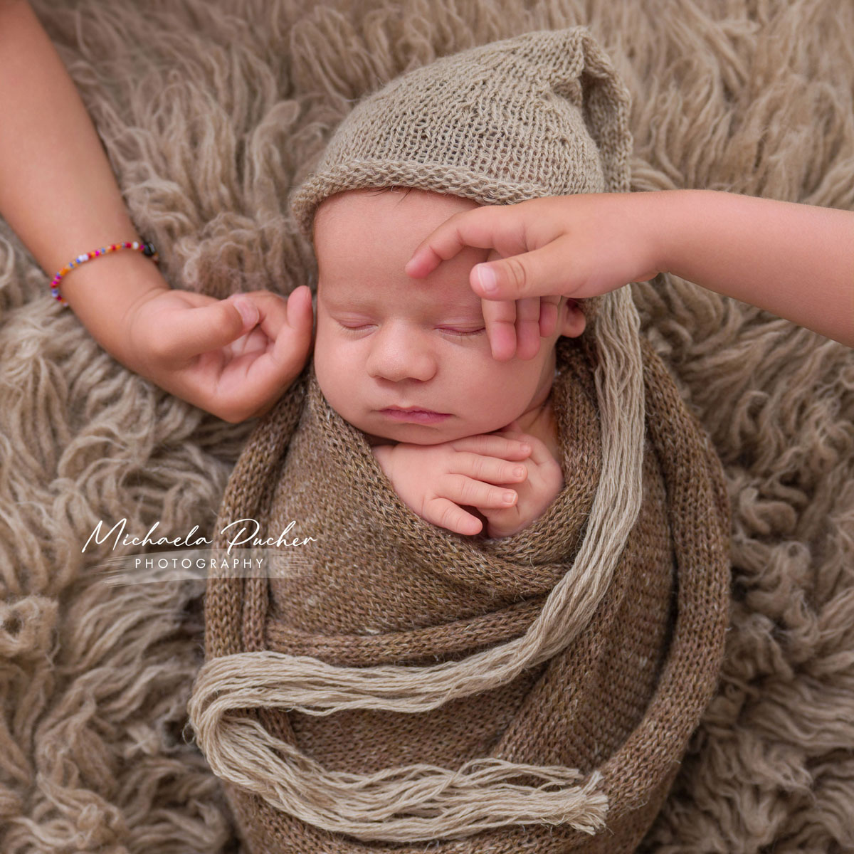 Schlafendes Neugeborenes in braunen Stricktüchern, während ein Geschwisterkind ihm sanft über die Stirn streicht.